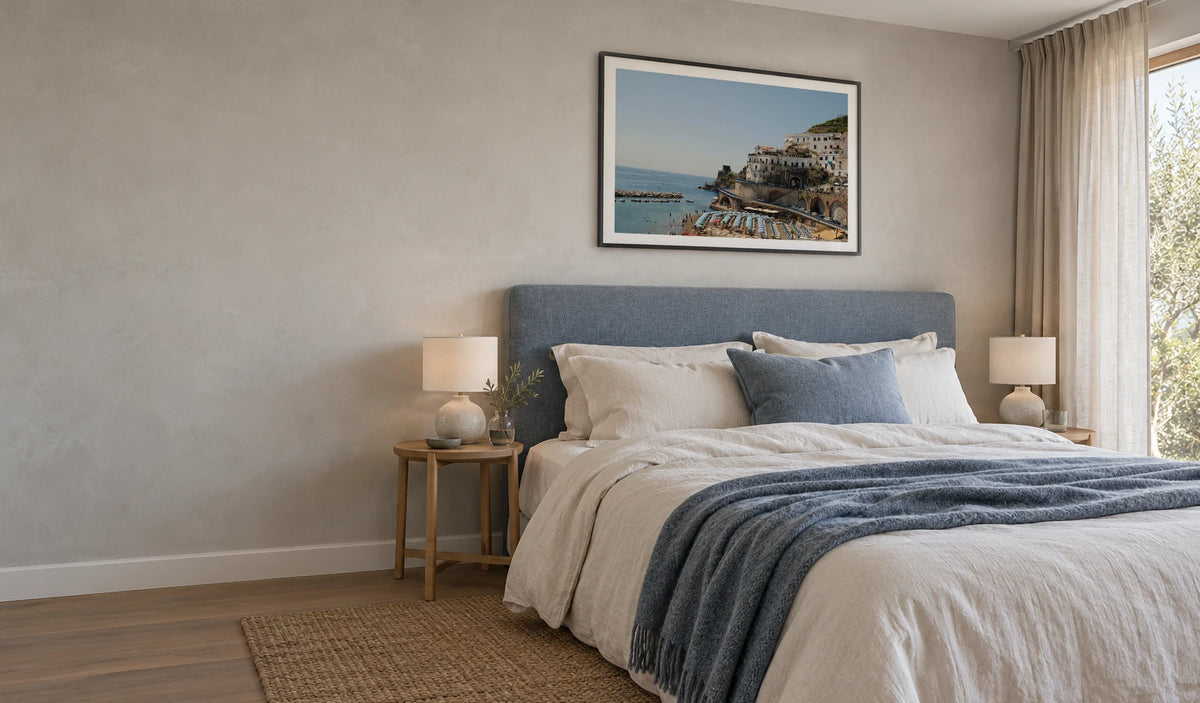 Amalfi Coast photograph in a black frame, hung above a blue upholstered bed with natural room perspective and daylight.
